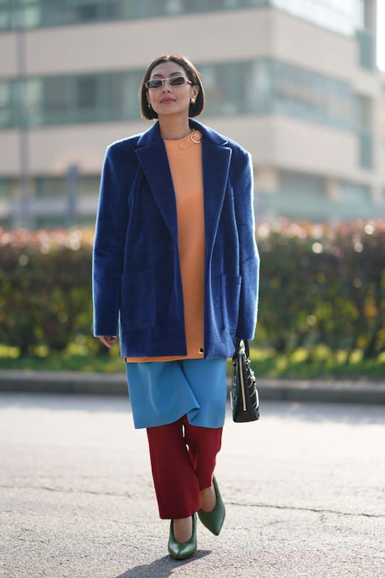 MILAN, ITALY - FEBRUARY 19: A guest wears sunglasses, earrings, a blue fluffy oversized jacket, an orange top, a blue skirt, red pants, green pointy shoes, a bag outside Gucci, during Milan Fashion Week Fall/Winter 2020-2021, on February 19, 2020 in Milan, Italy. (Photo by Edward Berthelot/Getty Images)