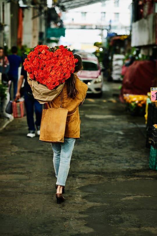 Wide shot rear view of woman carrying large heart shaped bouquet of roses over shoulder after shopping in flower market during vacation