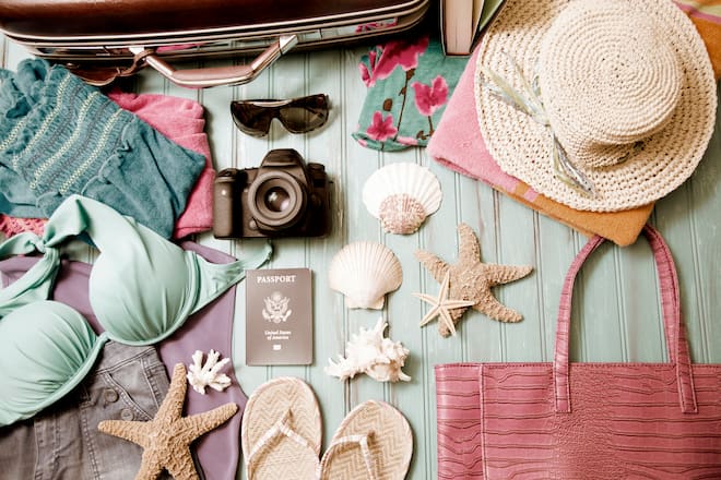 Summer vacation items ready for packing into suitcase. Items arranged in knolling pattern. No people. Items include: clothing, straw hat, binoculars, camera, flip flops, shoes, sea shells, book, suitcase, beach towel, sunglasses, passport.