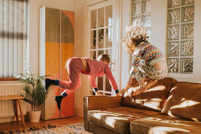 Two cute kids wearing pyjamas bounce on a brown leather couch in a sunny domestic room. Conceptual with space for copy.
