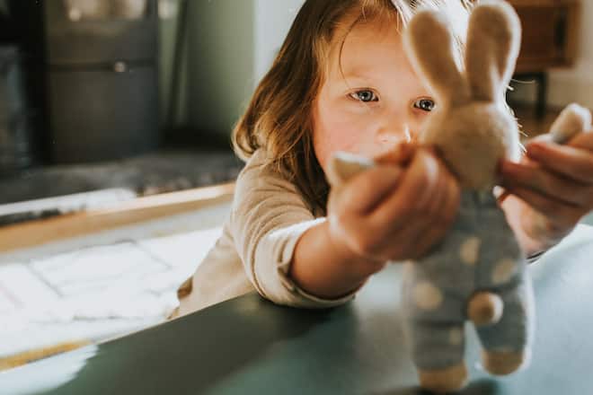 A curious child holds a small toy bunny rabbit and her expression is that of curiosity, suspicion, or intrigue as she looks intently at it. Domestic environment in the background provides space for copy.