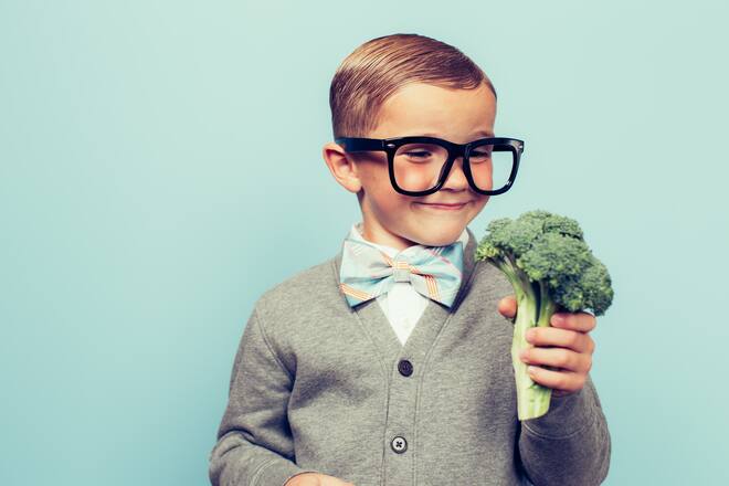 A young nerd boy with glasses is excited to eat his vegetables. He is dressed in bowtie and nice vest and glasses with a big smile at his broccoli.