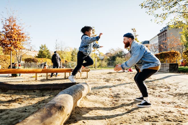 A young boy leaping into his fathers arms from a log while messing around in a playground at the park together.