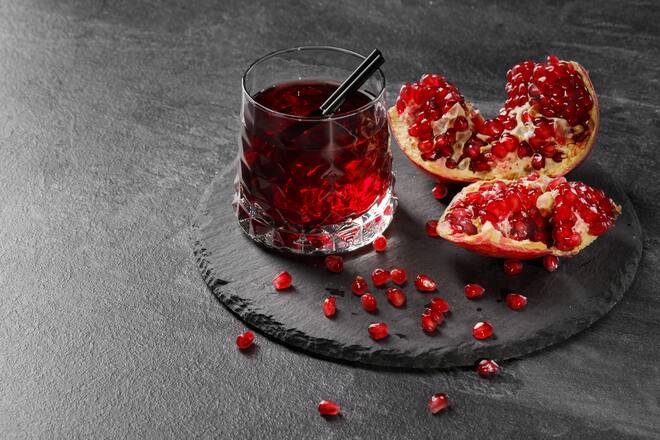 A transparent glass full of sweet pomegranate juice and a cut garnet on a round plate on a dark gray background. Summer, healthful and fruity red cocktail with black straw and fresh garnet.