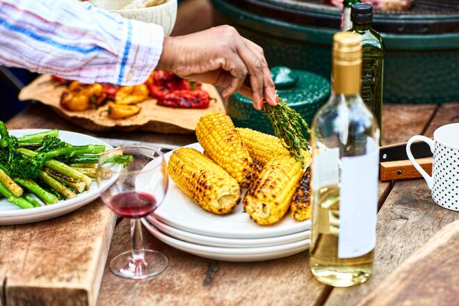 Char grilled corn with green vegetables and wine on table, woman seasoning food with herbs, food preparation, healthy eating